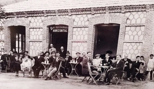 Fotografía antigua en blanco y negro: un grupo numeroso posa sentado ante la fachada de ladrillo del «Bar Restaurant Horizontal»