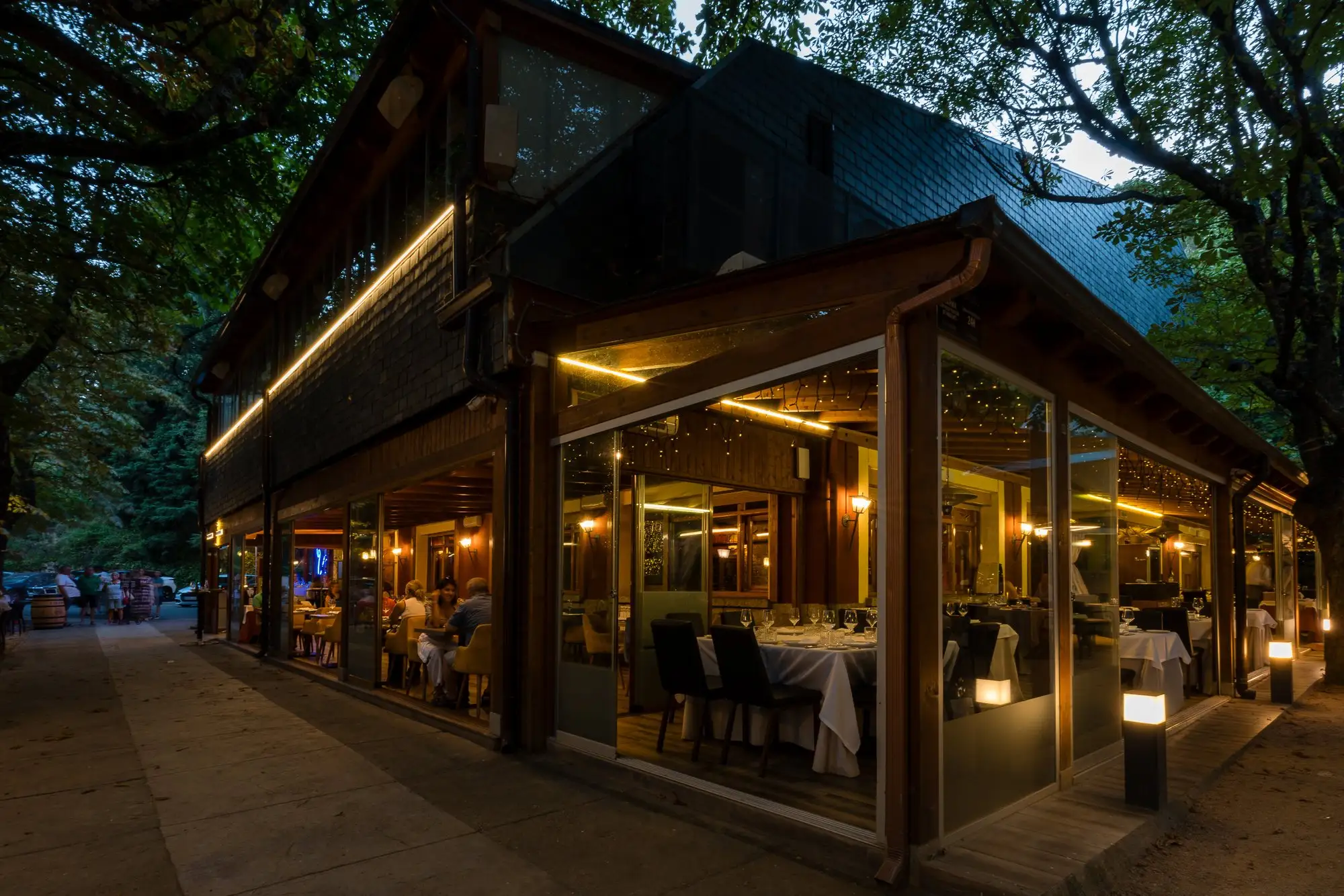 El comedor acristalado de noche visto desde fuera, con las mesas vestidas y las luces encendidas bajo los árboles