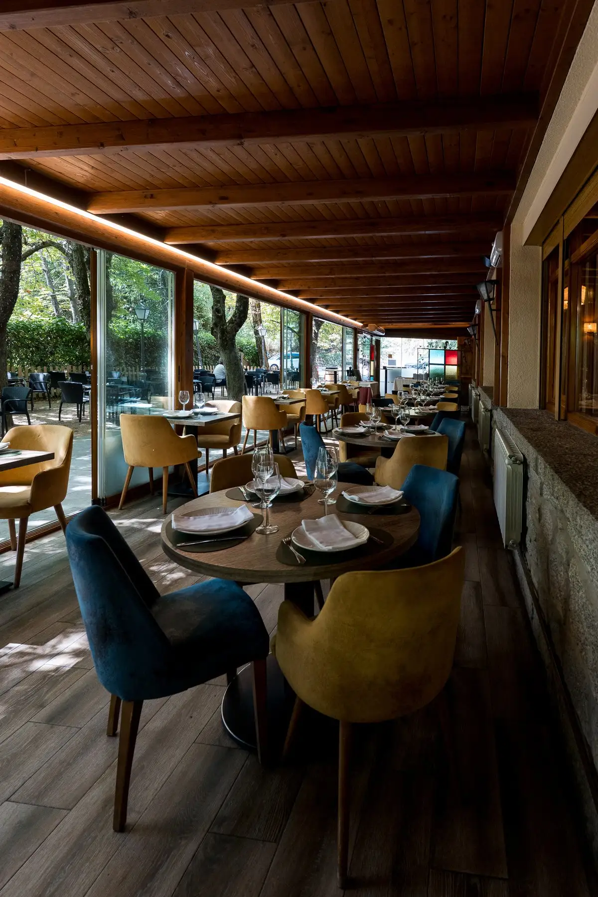 Comedor acristalado con techo de madera, mesas vestidas y sillas tapizadas en mostaza y azul, con la terraza al otro lado del cristal