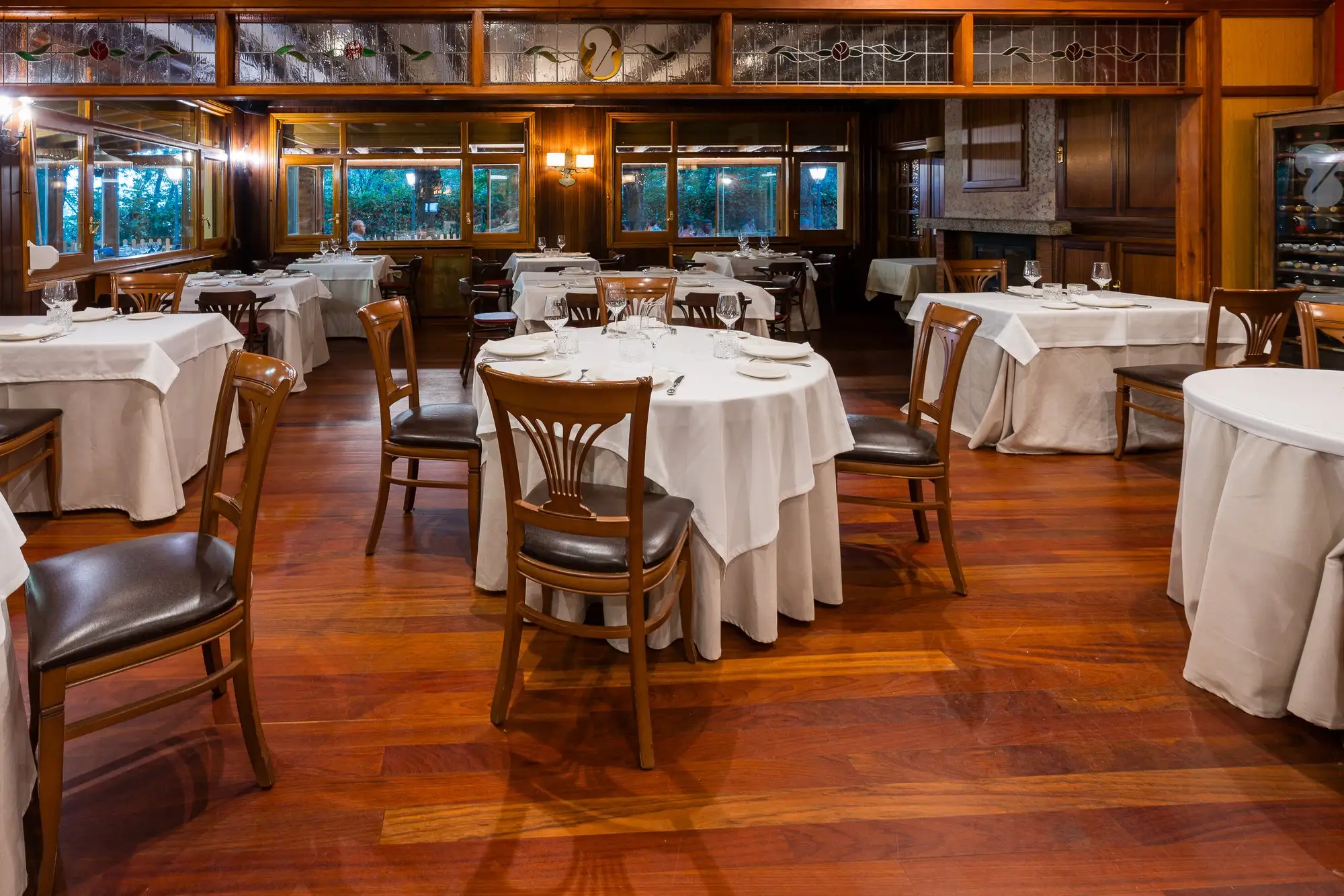 Comedor de suelo y paredes de madera, con mesas redondas de mantel blanco y ventanales al bosque