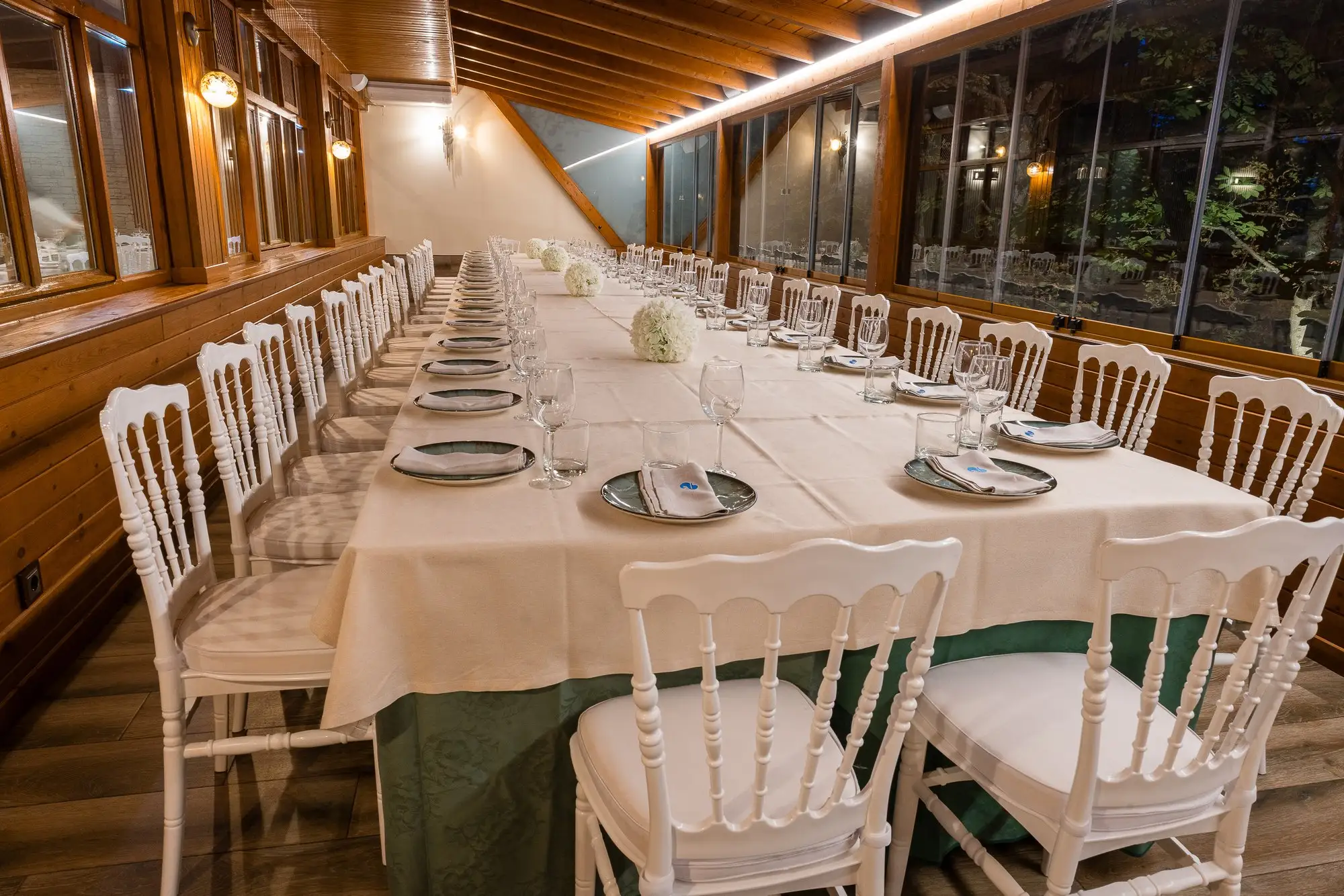 Mesa larga vestida para una celebración en el comedor acristalado, con sillas blancas y centros de flores blancas