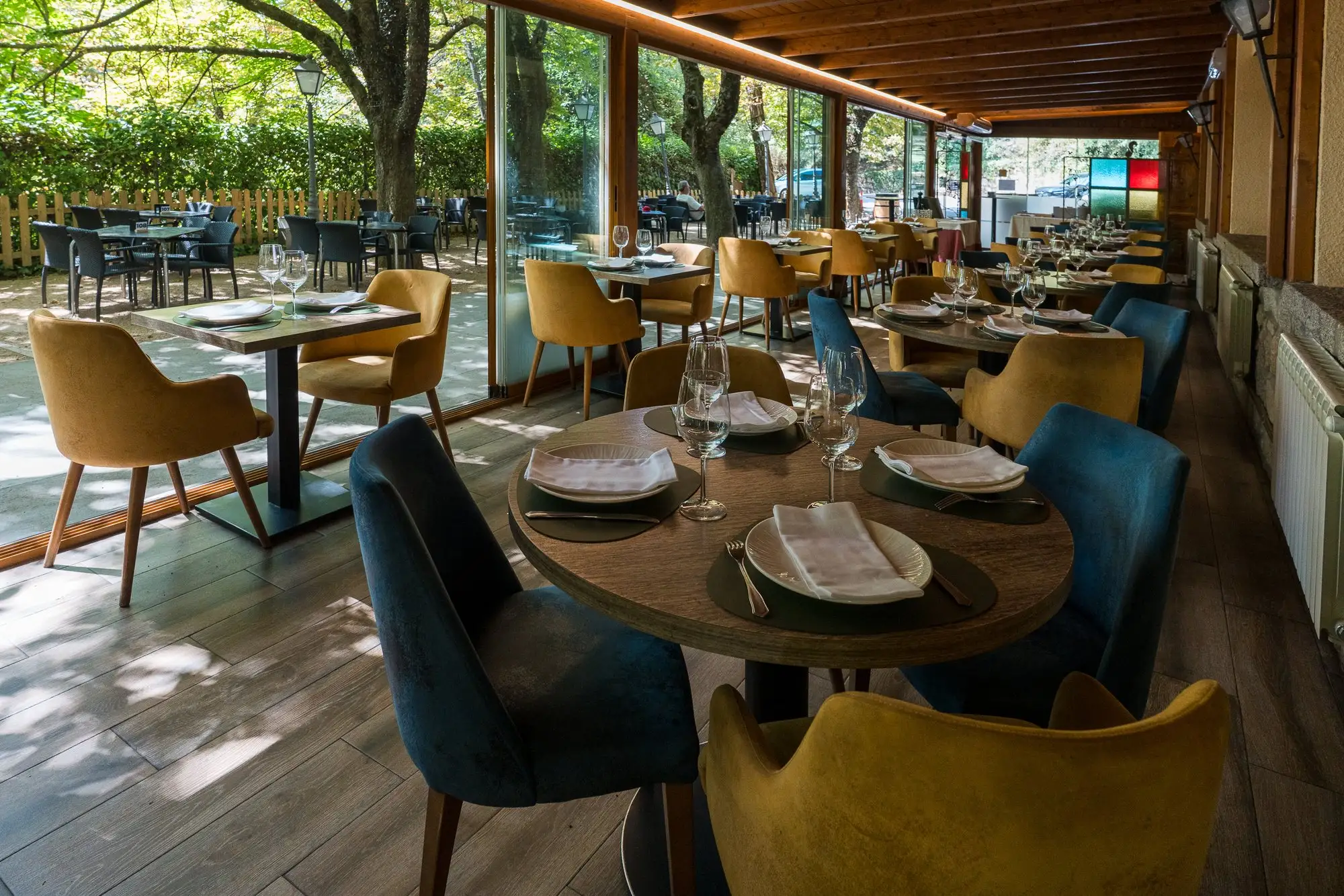 Porche acristalado con mesas de madera y sillas tapizadas en mostaza y azul, abierto a la terraza y a los árboles