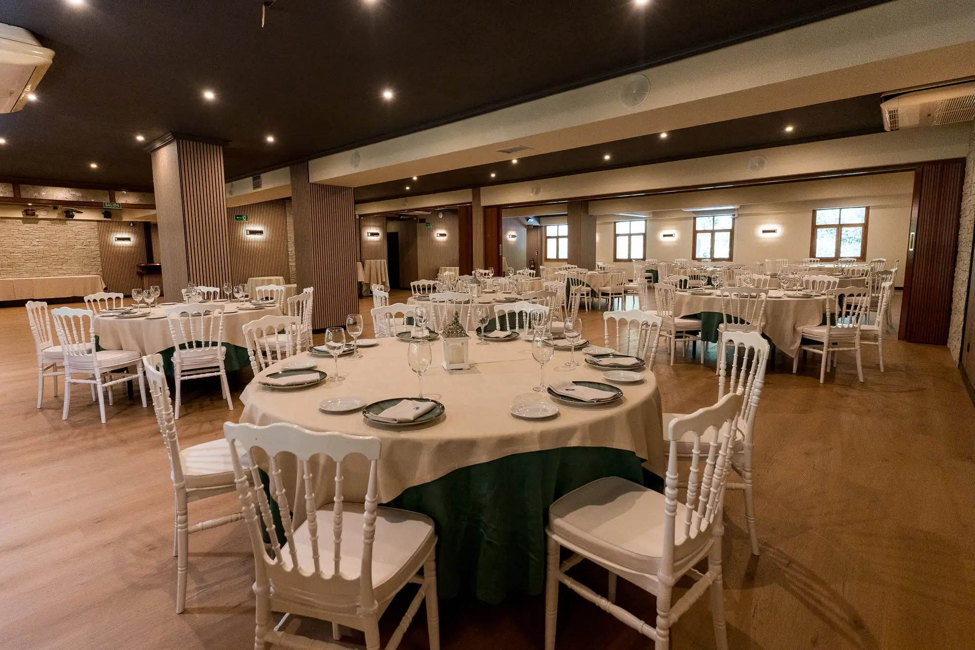 Salón de celebraciones montado con mesas redondas de mantel verde y sillas blancas