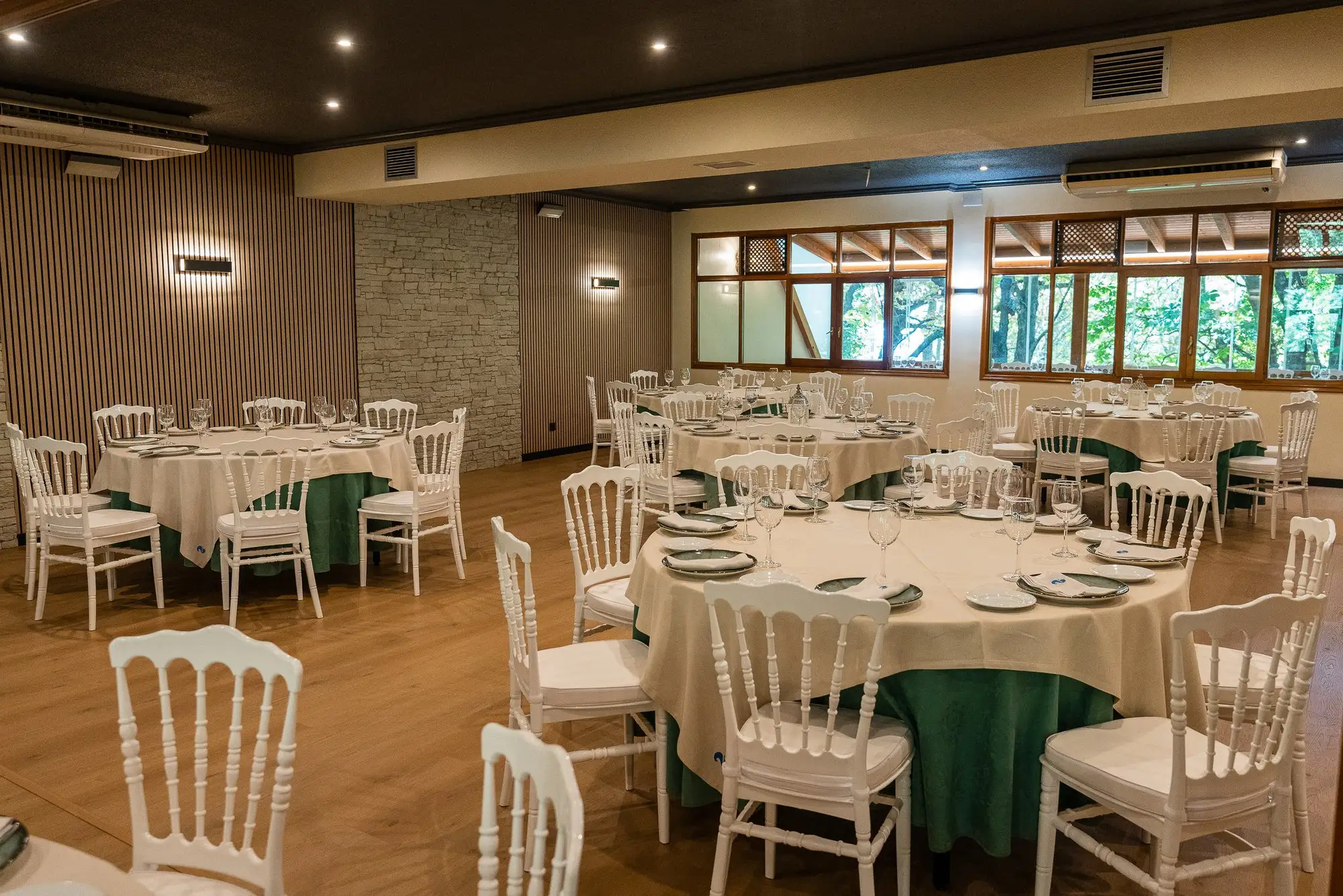Salón de celebraciones con mesas redondas en verde y crema, sillas blancas y ventanales al bosque