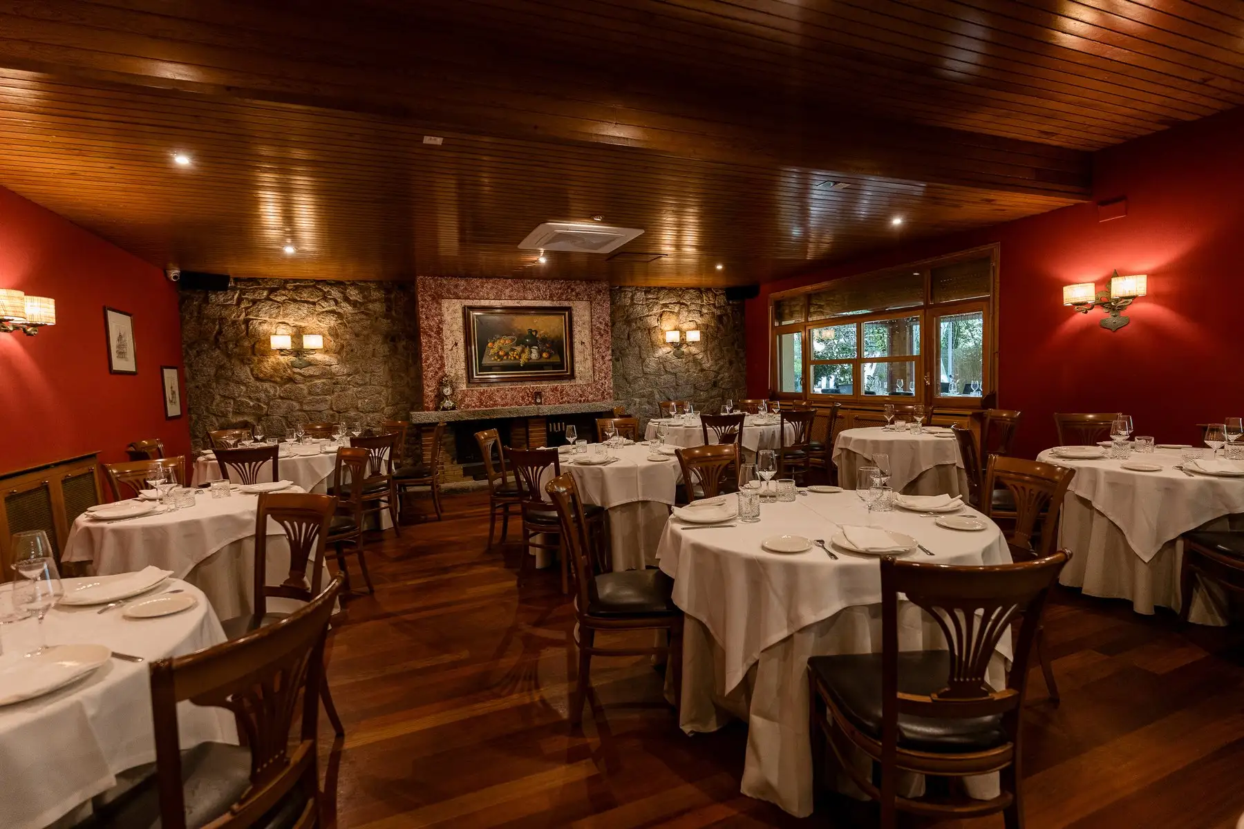 Salón interior de paredes rojas y techo de madera, con mesas redondas vestidas y una chimenea al fondo, entre muros de piedra