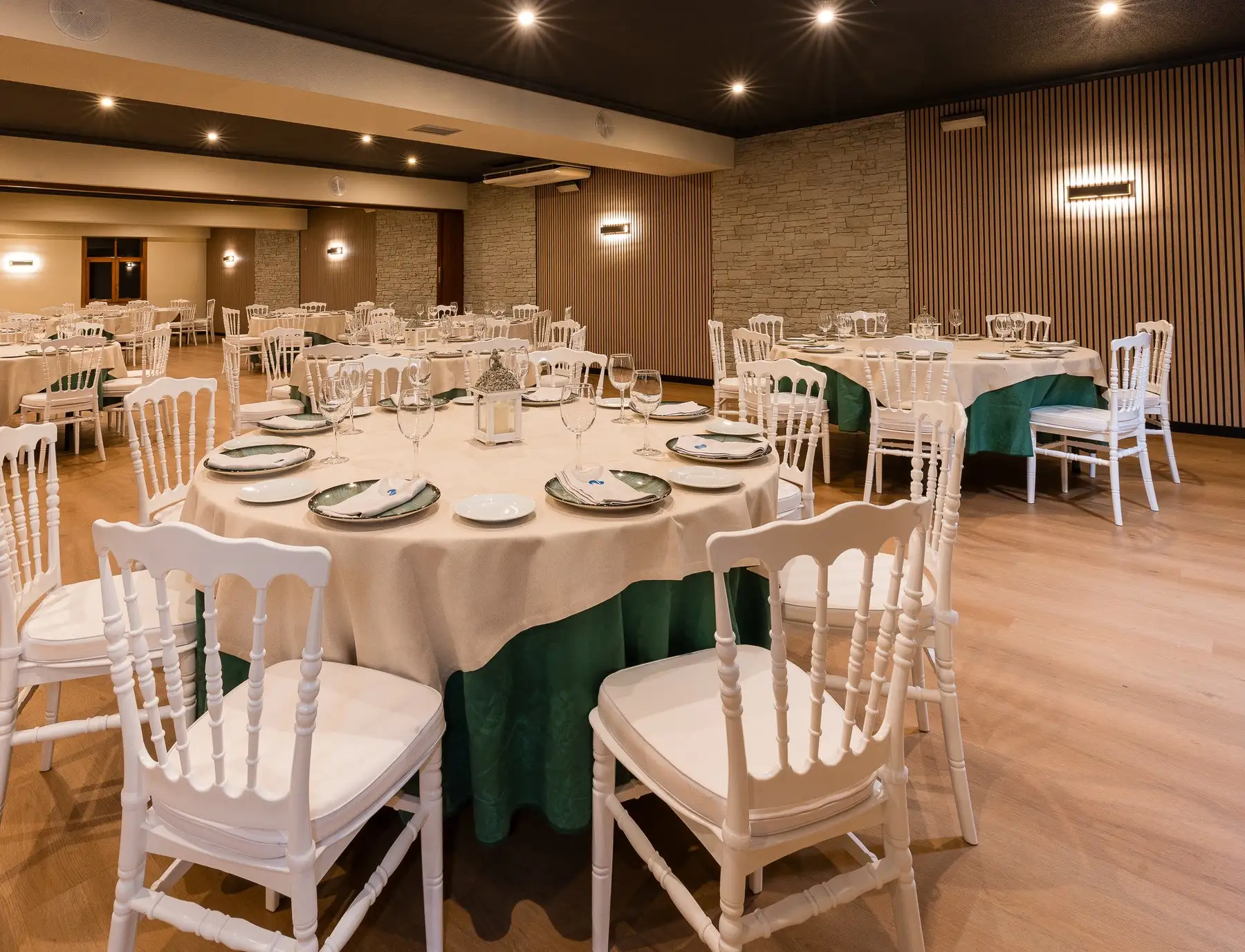 Salón de eventos con mesas redondas vestidas en verde y crema, sillas blancas y pared de piedra