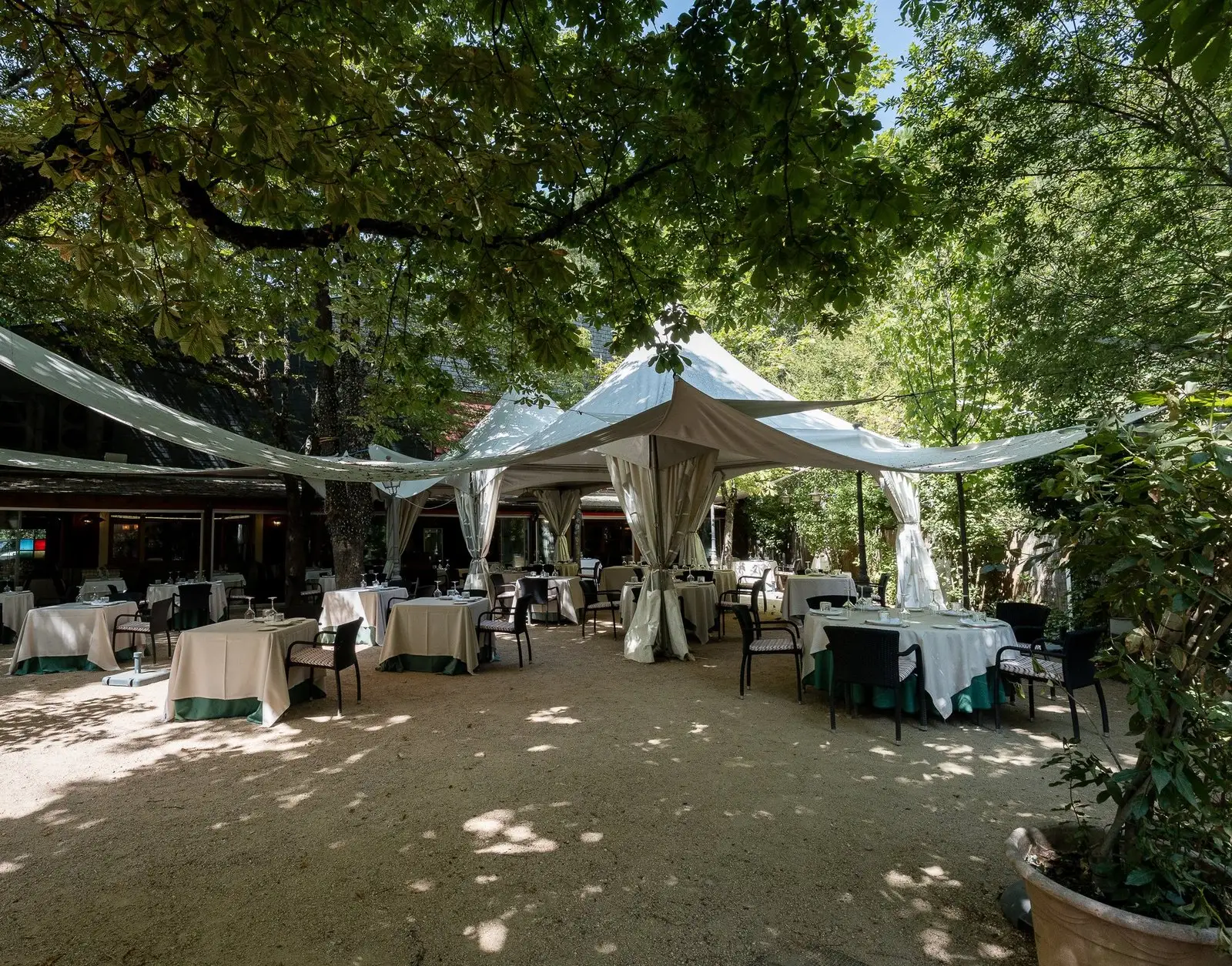 La terraza de Restaurante Horizontal de día: mesas vestidas al aire libre bajo los árboles y las carpas blancas, con el edificio al fondo