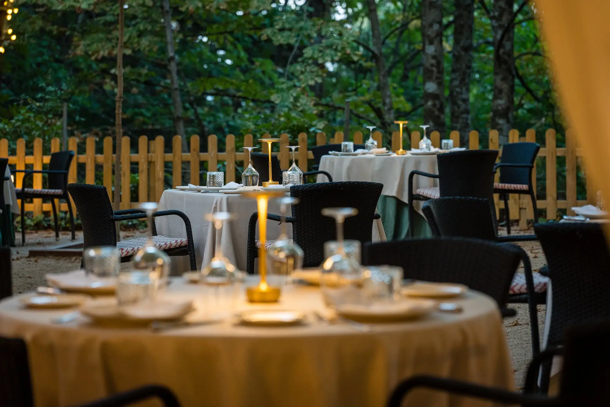 Mesa redonda de la terraza al anochecer con su lámpara encendida, junto a la valla de madera y el bosque