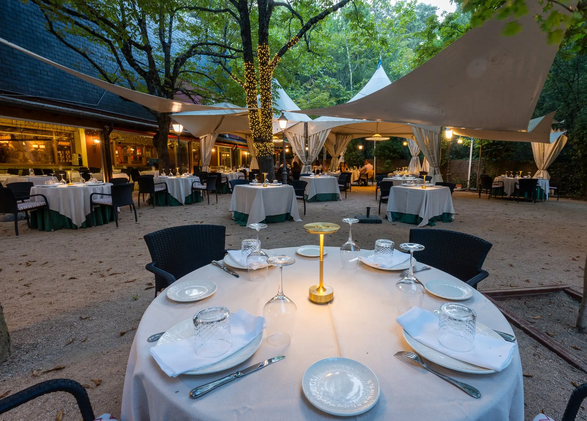 Mesa redonda vestida con una lámpara encendida en primer plano y la terraza con sus carpas al fondo