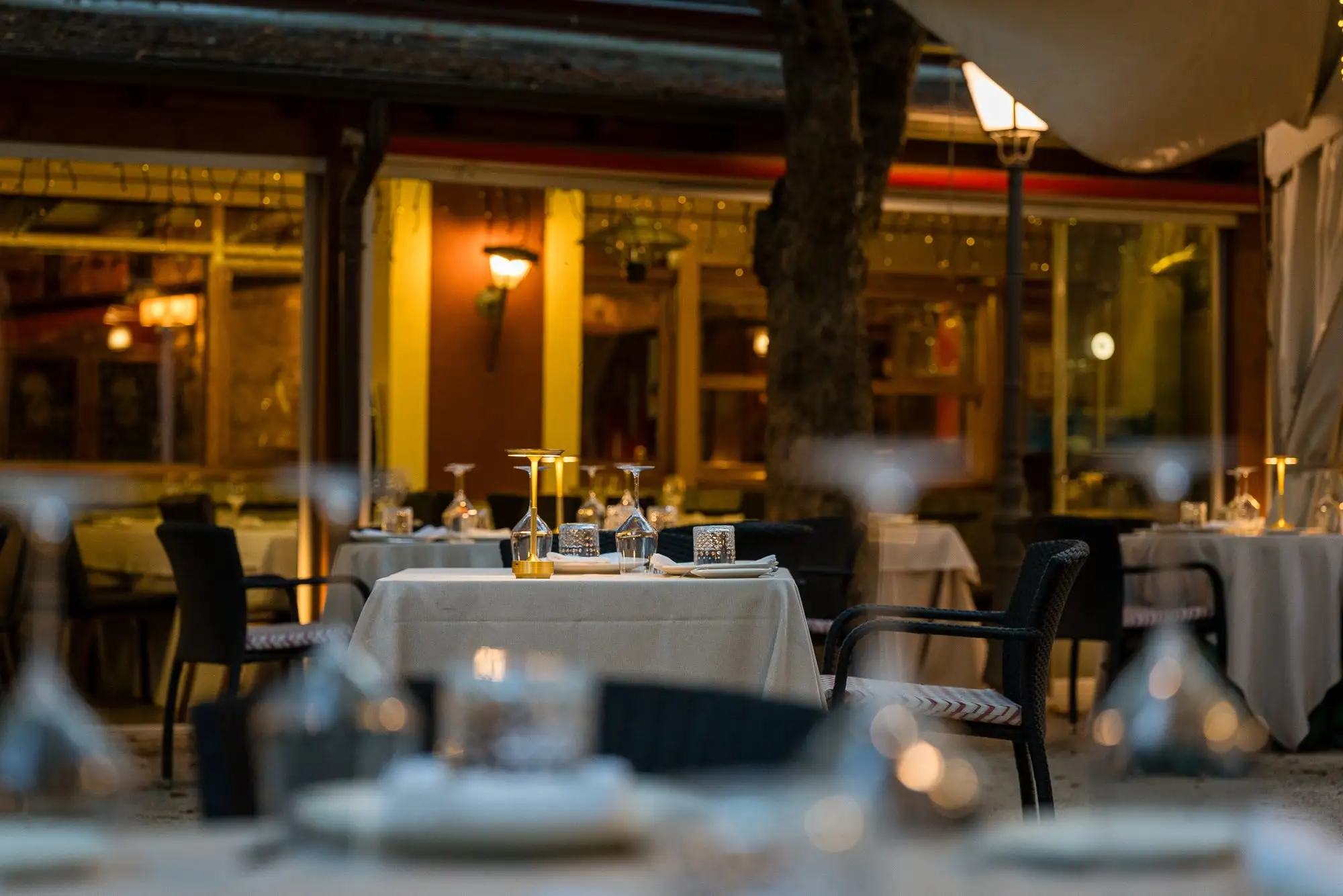 Mesas de la terraza por la noche, con las copas en primer plano y los ventanales iluminados del restaurante detrás